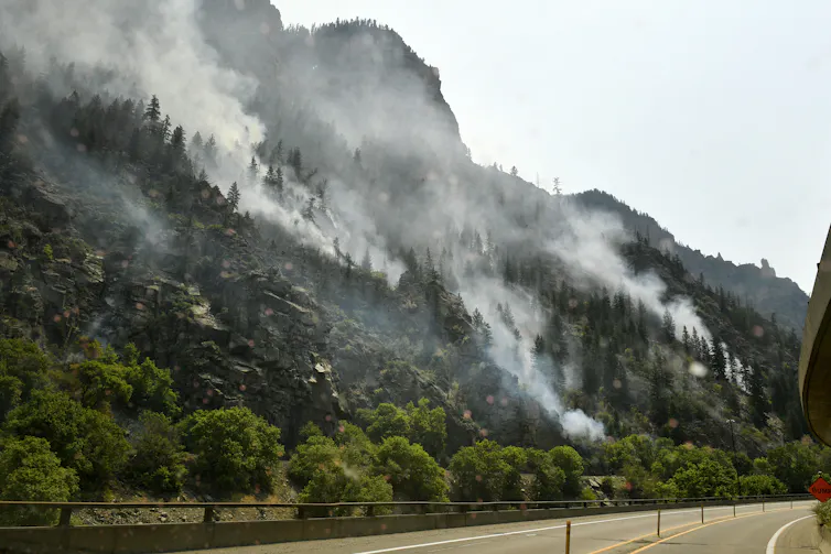 Smoke billows from a rocky and mountainous forest near an empty highway.