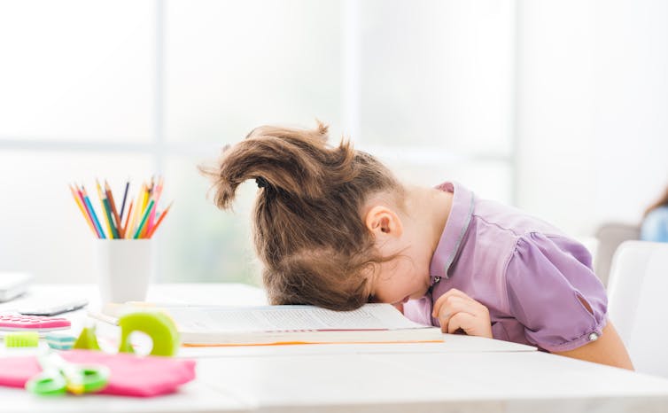 a young girl sitting at a desk buries her forehead in a textbook