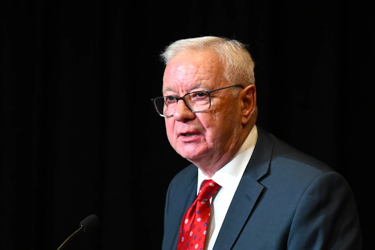 A man with white hair and glasses against a black backdrop