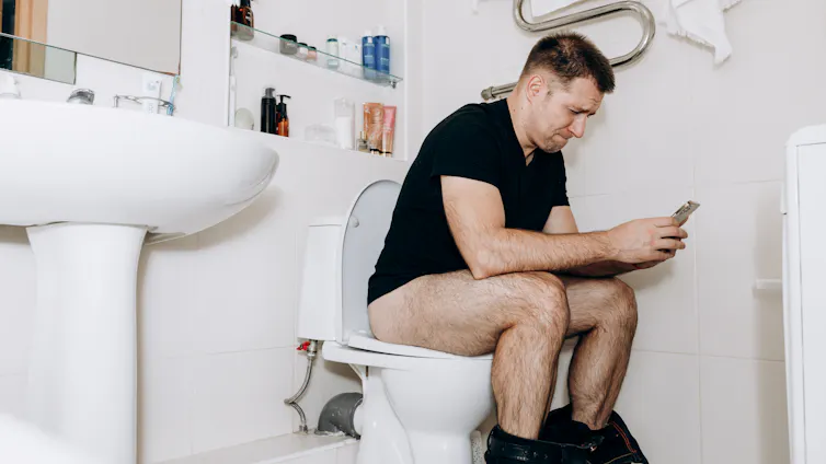 A man sits on the toilet using his smartphone. He looks frustrated.