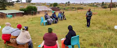 A man stands up and talks with several people who are sitting in a circle on chairs. Everyone is sitting outside on open green grasslands with a rural village in the background.