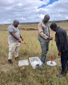 Three people stand outside in brownish grass on a huge open grassland measuring and taking samples of the grass and soil.