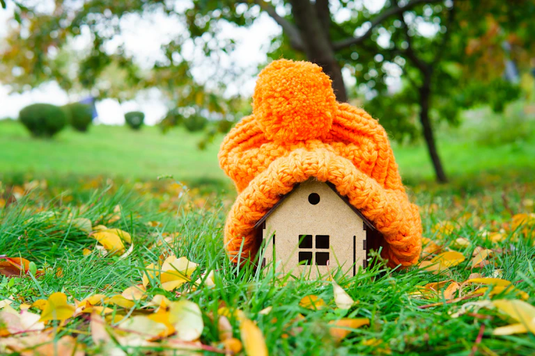 Chunky winter hat put on a wooden house model placed on a green lawn with autumn leaves in countryside
