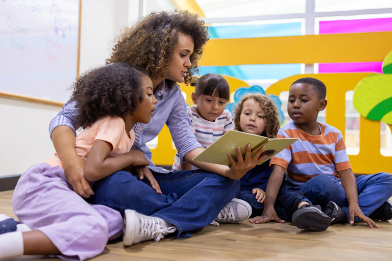 A young woman with curly hair reads to four children sitting against her on the floor.