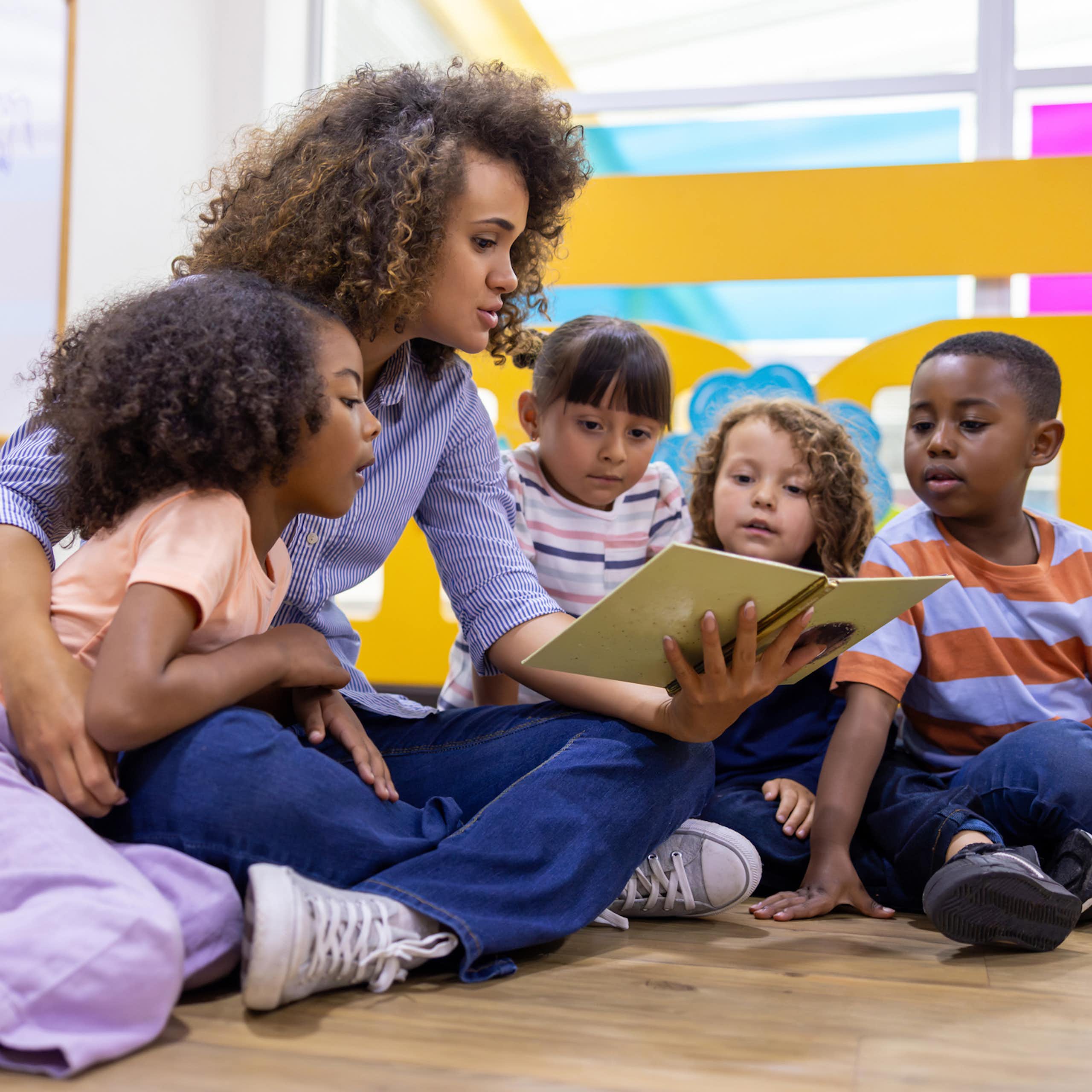 A young woman with curly hair reads to four children sitting against her on the floor.