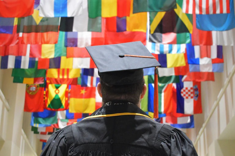 A student wearing a black robe and graduation cap is seen from behind, walking toward many country flags hanging from a ceiling.