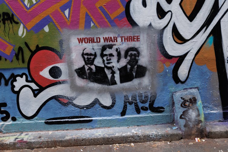 A wall has graffiti on it including a stencil of three men under the words 'World War Three.'