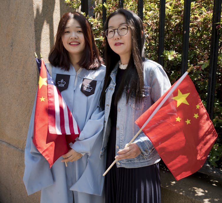 Two young Chinese women with dark hair hold red flags with yellow stars on them. One of the women wears a light blue graduation robe and smiles.