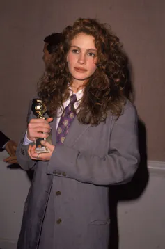 Young woman with long, curly, brown hair holds an award while wearing a bulky, gray jacket and a purple tie.