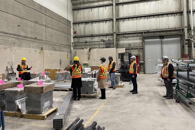 Workers in orange vests at hard hats stand inside warehouse at a construction site.