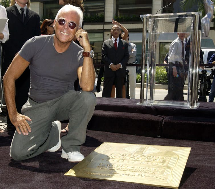 Tanned man with white hair wearing sunglasses poses on one knee next to a gold plaque.