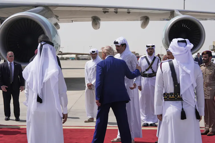 A rotund man with fluffy white-blond hair in a blue suit is photographed from behind on a miltary base tarmac speaking to a man wearing a white robe and a keffiyeh.