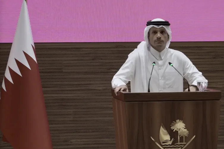 A man in a white robe and wearing a keffiyeh stands behind a podium in front of a wall painted pink.