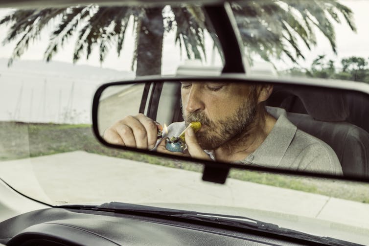 View of a man's reflection as he smokes marijuana through a pipe at the wheel of a car.