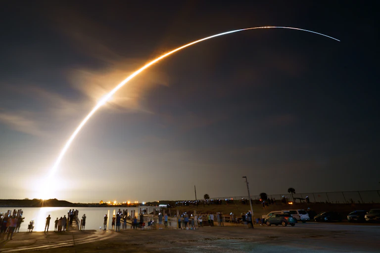 A group of people on a beach watching a space launch which lights up the sky in a bright arc.