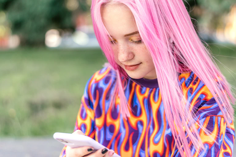 Une jeune fille aux longs cheveux rose, portant un pull de couleurs vive, regarde son smartphone.