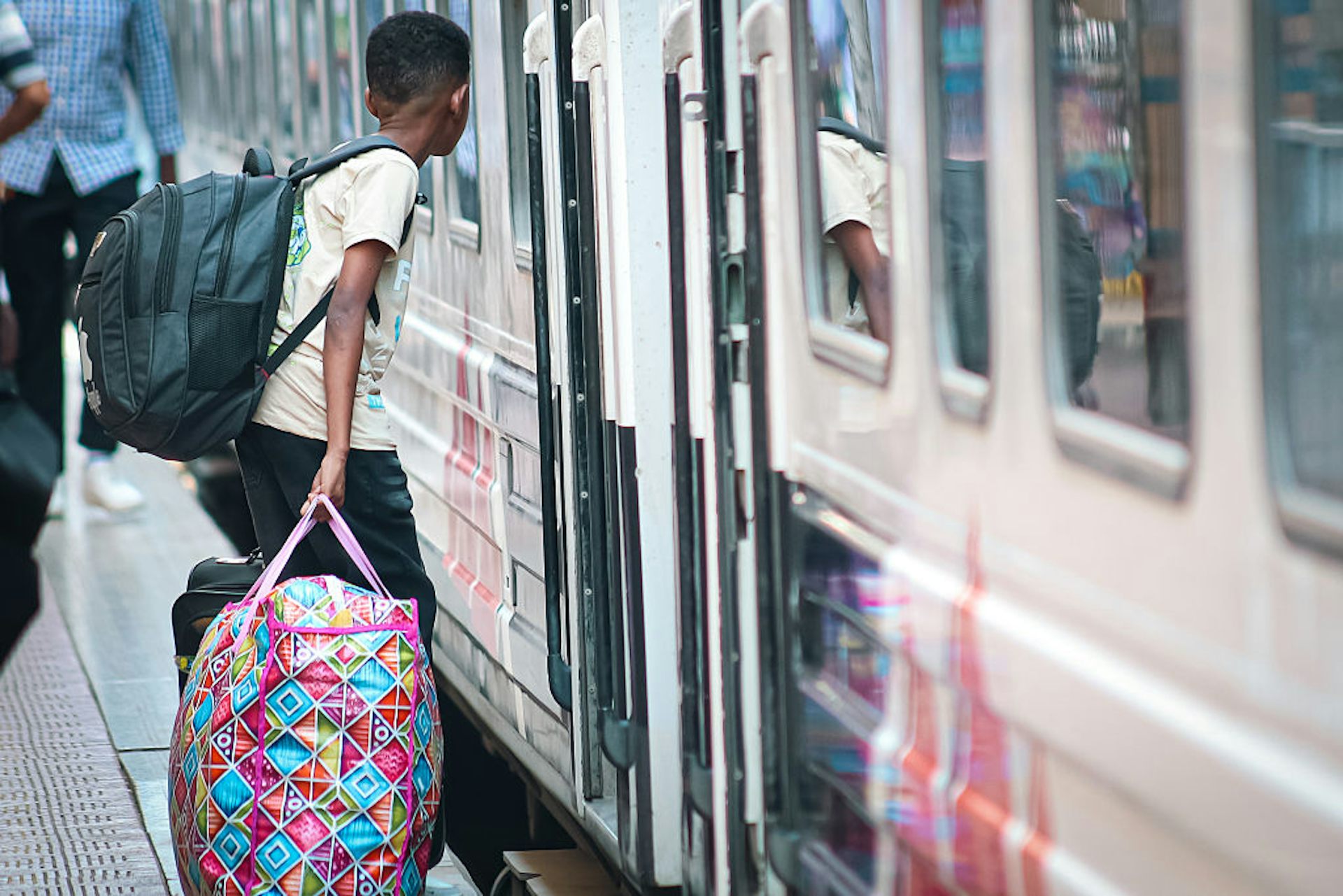 A boy carrying a large bag in one hand and a packed backpack walking into the doorway of a train with a white exterior. 