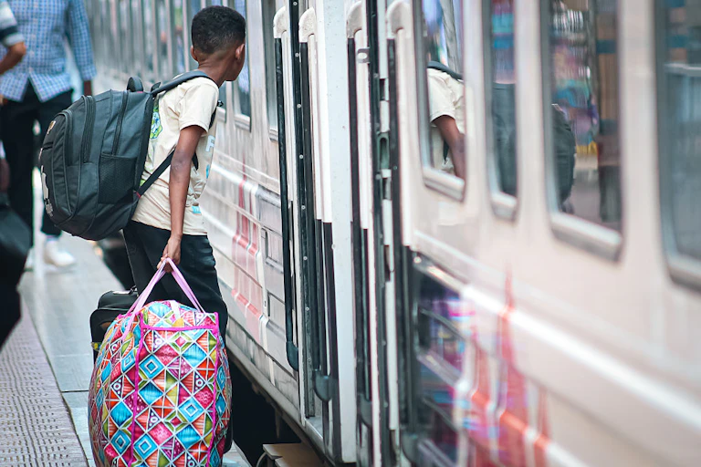 A boy carrying a large bag in one hand and a packed backpack walking into the doorway of a train with a white exterior.