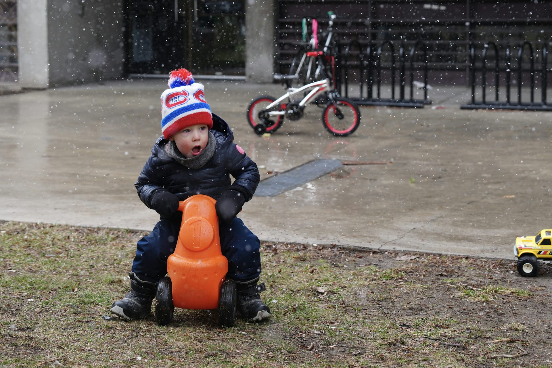 A child in a tuque and snowsuit riding on a small orange toy outdoors.