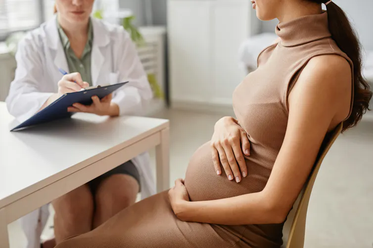 Cropped photo of a pregnant woman in a beige dress sitting at a table talking to a health-care worker