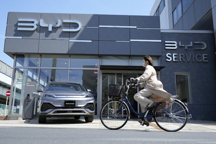 A woman rides a bicycle past a car dealership