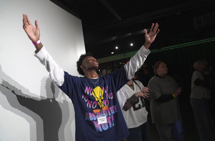 A man wearing a long-sleeved t-shirt raises his arms in prayer inside a dark room.
