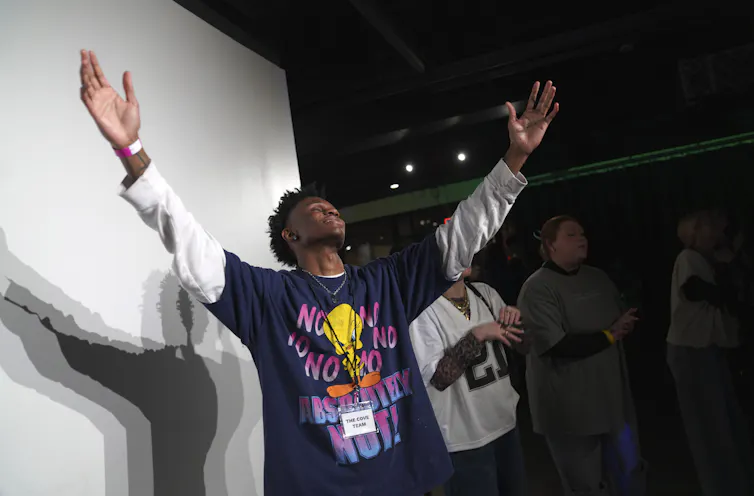A man wearing a long-sleeved t-shirt raises his arms in prayer inside a dark room.