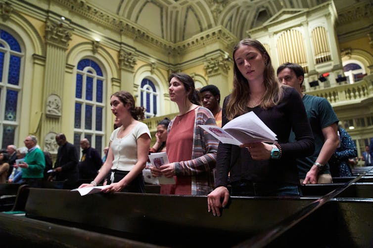 Three young women stand in church pews in a lofty sanctuary as they pray or sing.