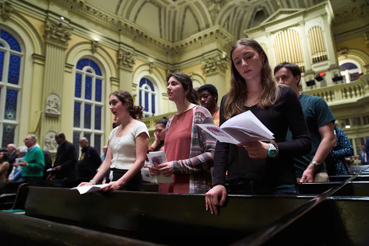 Three young women stand in church pews in a lofty sanctuary as they pray or sing.