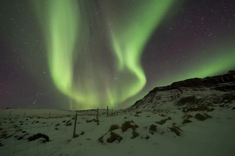 Green lines of light crossing the night sky, above a snow-covered landscape.