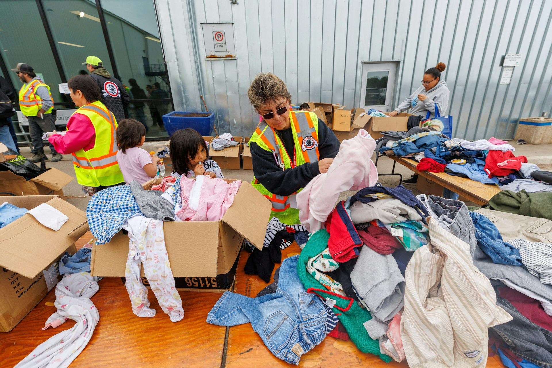 A woman and young girl stand behind a pile of clothes on a table. The woman holds up an item of clothing. 