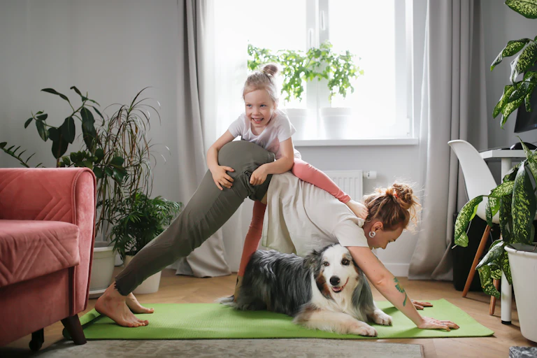 Laughing kid climbs on a woman doing a yoga pose at home with their dog.