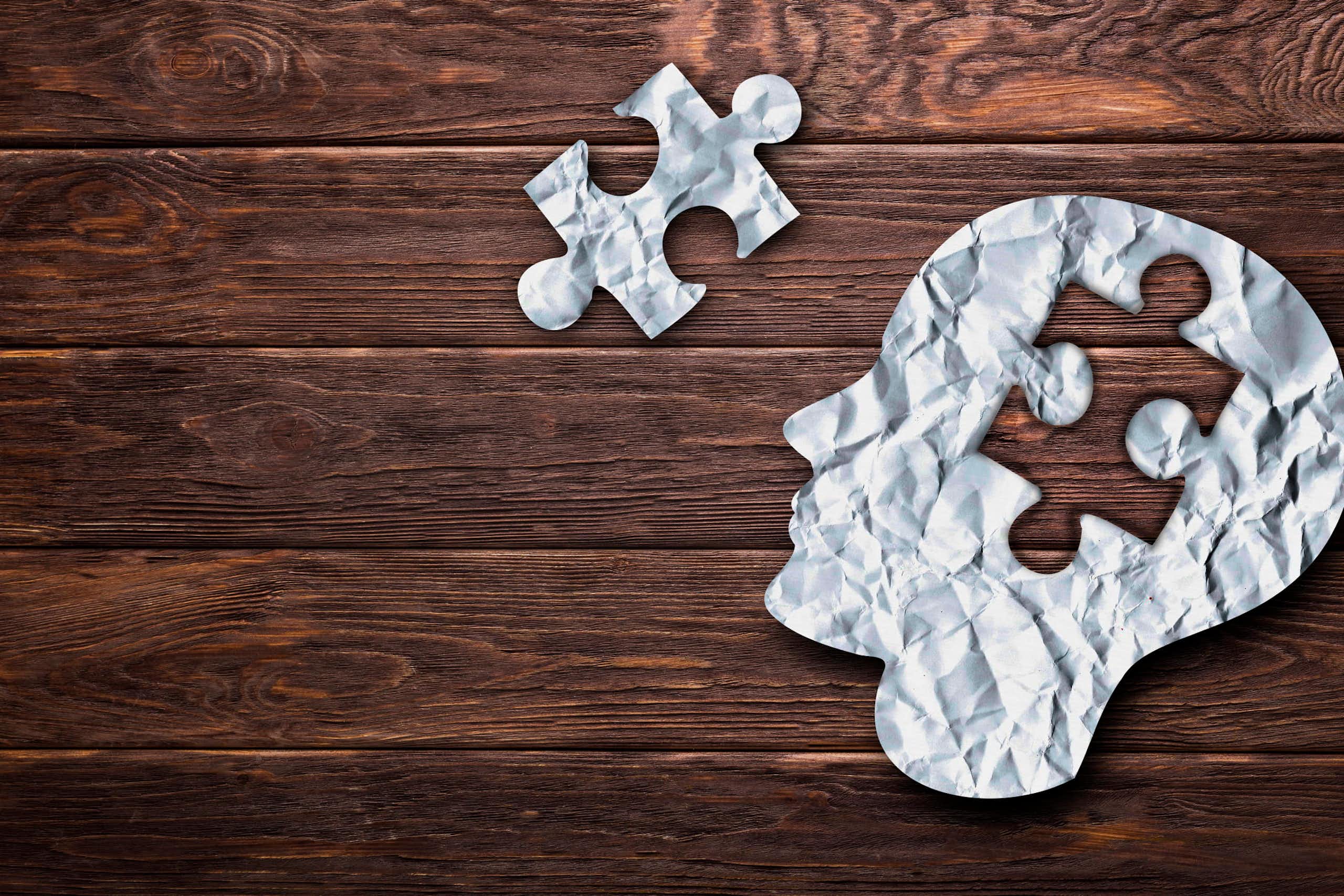 Paper silhouette of a head with a puzzle piece cut out of it against a wood plank background