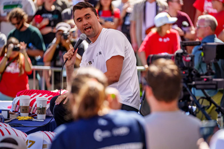 A young white man with dark hair and a white shirt speaks into a microphone amid a crowd of people watching him.