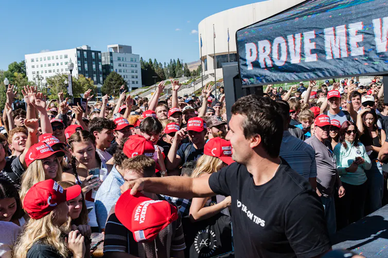 A young man wearing a black t-shirt extends his arm toward a crowd of young people, many of whom are wearing red hats.