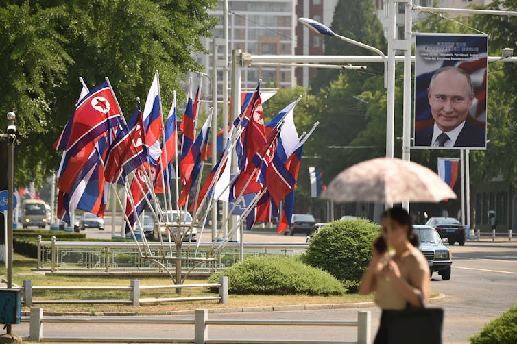 A woman with an umbrella walks past red and blue flags and a portrait of a man.