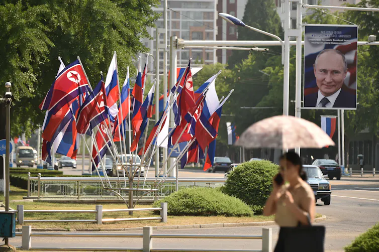 A woman with an umbrella walks past red and blue flags and a portrait of a man.