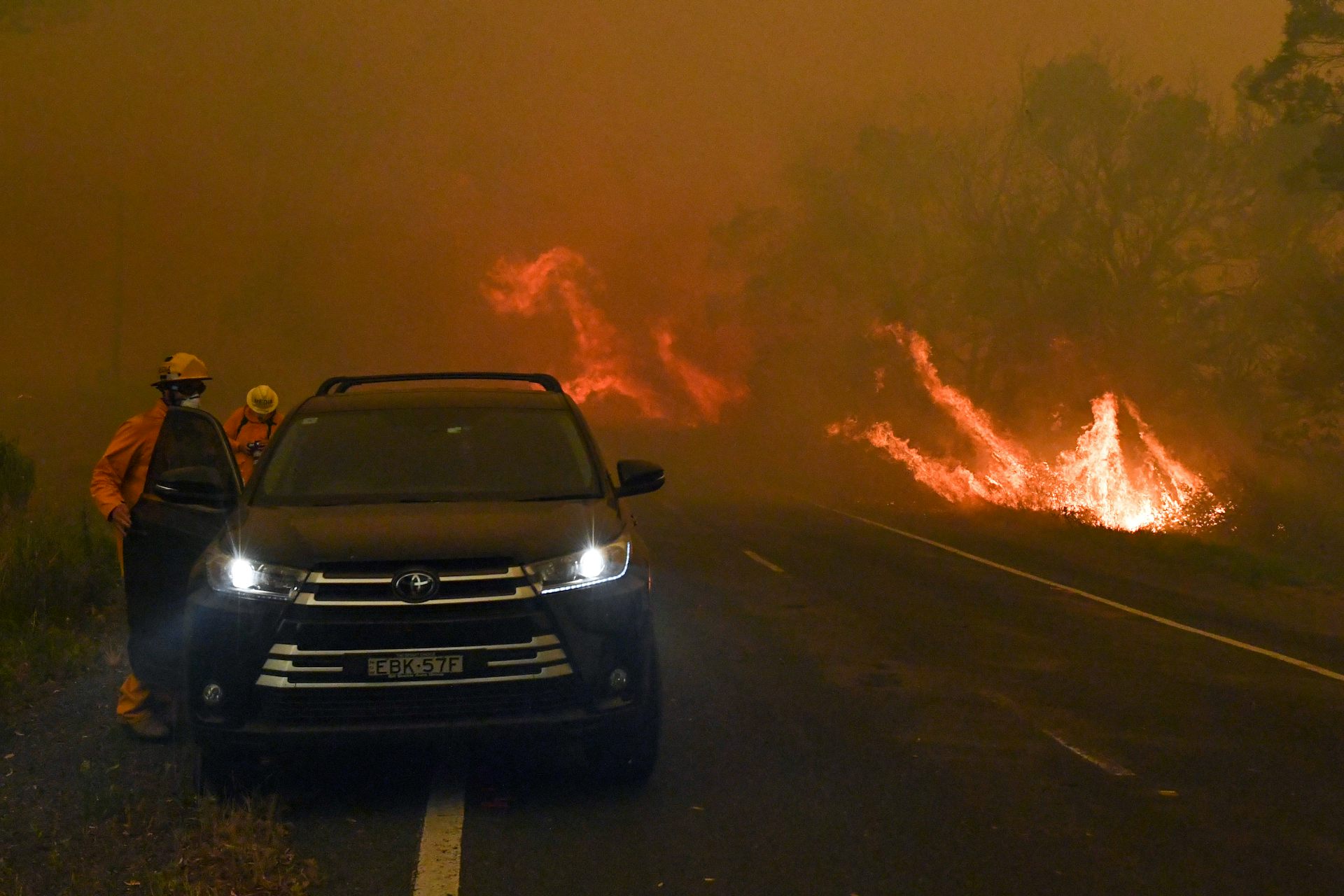 people stand near car as flames approach