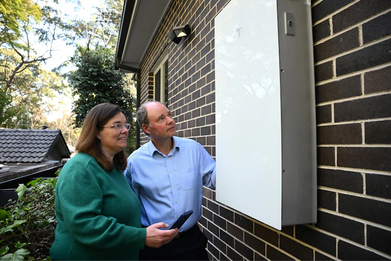 A middle aged couple standing by the side of their home, looking at their new home battery.