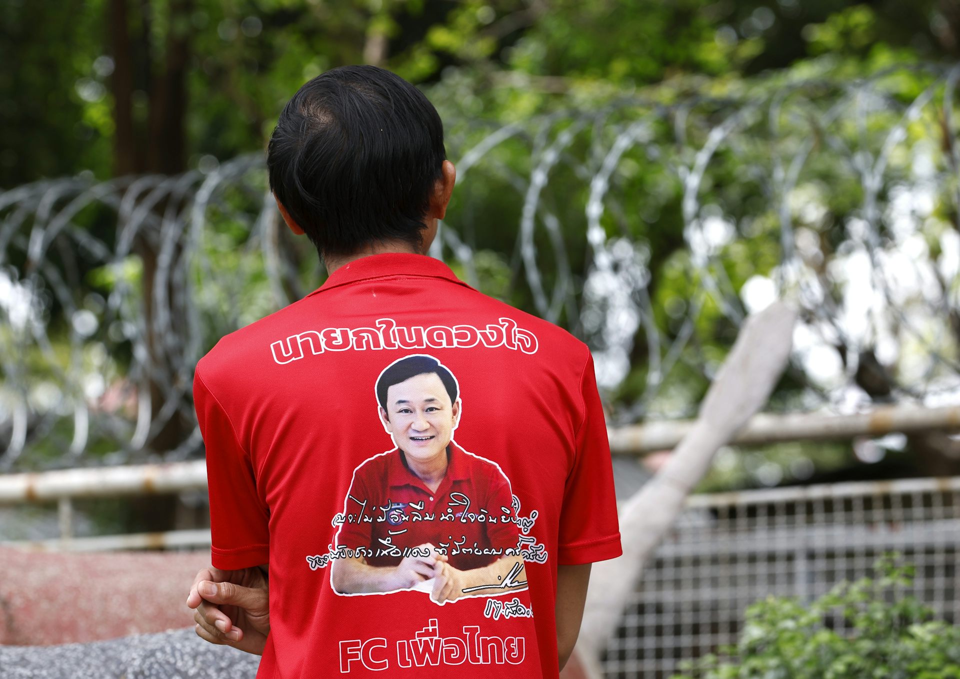 A man in a red t-shirt depicting Thaksin Shinawatra's face stands outside a prison.