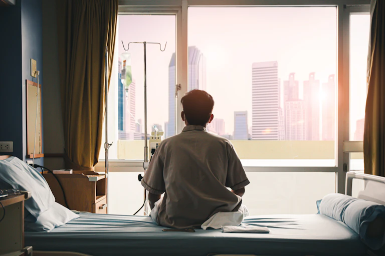 A hospital patient sits on his bed, looking out the window.