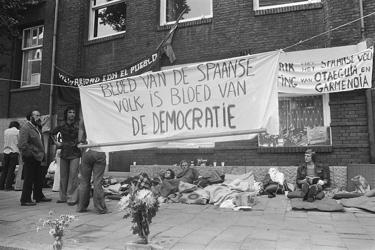 Foto en blanco y negro donde se ven varias pancartas en neerlandés y varias personas tumbadas con mantas en el suelo.