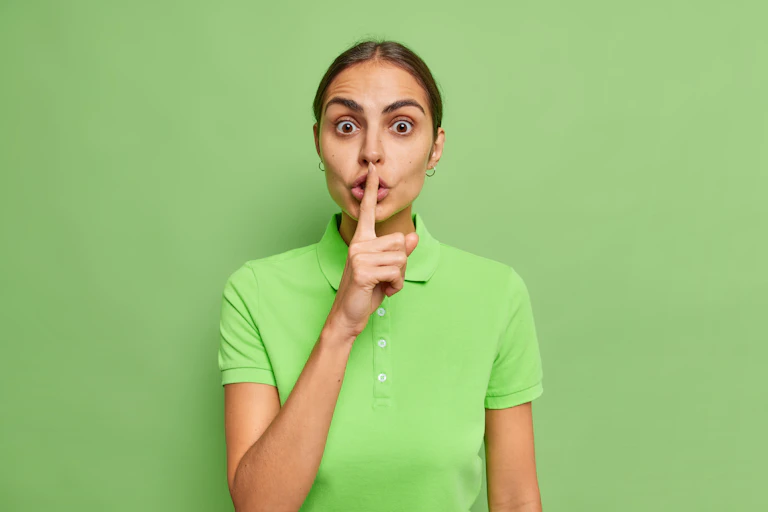 stock image of a woman in a green top against a green background holding her finger to her lips.