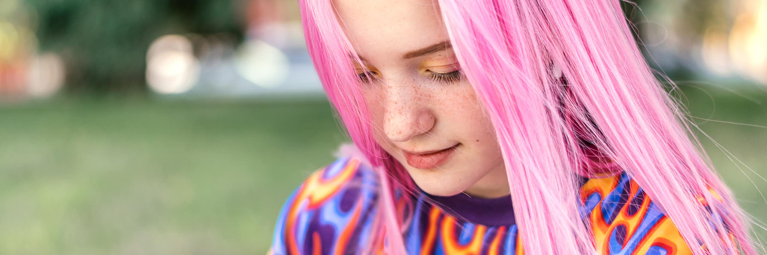 A young girl with long pink hair, wearing a brightly coloured jumper, looks at her smartphone.