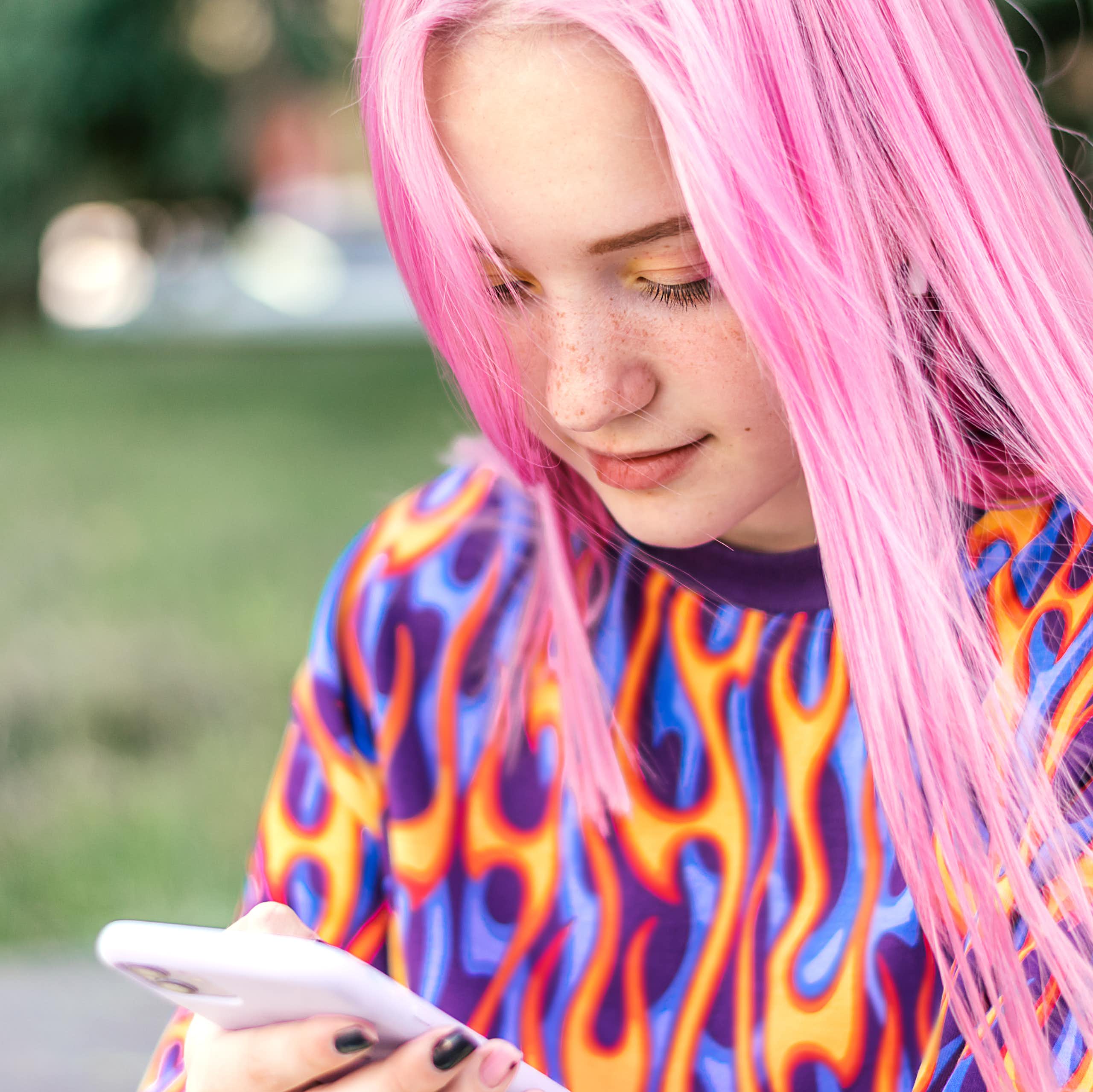 A young girl with long pink hair, wearing a brightly coloured jumper, looks at her smartphone.