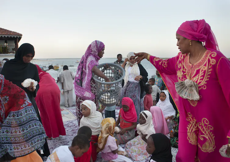 A woman in a bright pink dress and scarf hands something out as a woman passes with a basket and children sit on the ground.