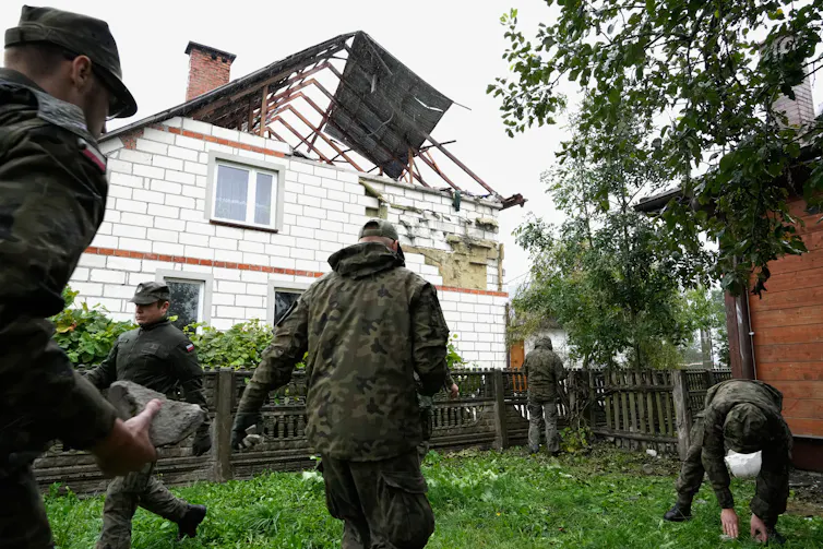 Workers pick up debris with a house in the background with a destroyed roof.