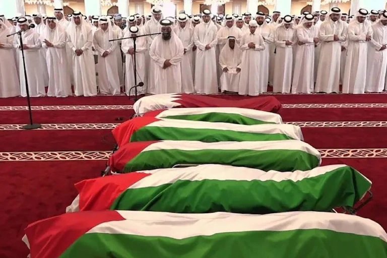 A line of Qatari men in traditional white clothing stand with their hands clasped looking at a row of coffins.