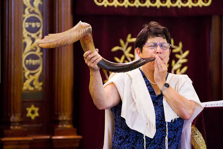 A woman in a blue dress and white prayer shawl blows into a large animal horn.