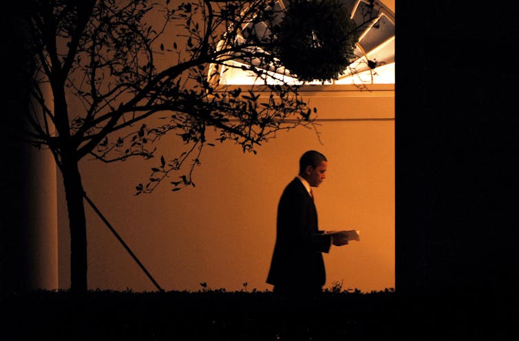 A man in a suit walking on an outside path next to a white building bathed in orange light.